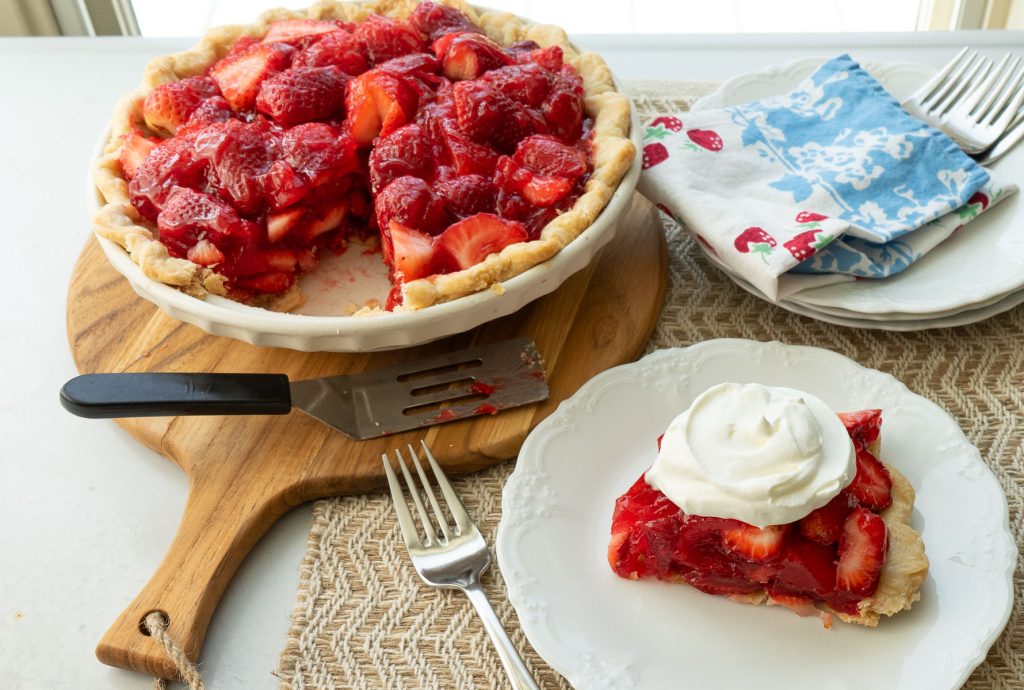 fresh strawberry pie with once slice taken out, a slice of strawberry pie on a white plate with whipped topping and a fork laying to the left of the plate