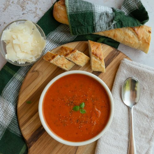 bowl of roasted tomato basil soup with a bowl of parmesan in upper left hand corner of the photo and a baguette wrapped in a kitchen towel in top of photo. spoon on tan napkin to the right of the bowl. soup is garnished with pepper and basil leaves