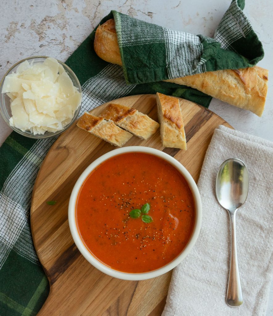 bowl of roasted tomato basil soup with a bowl of parmesan in upper left hand corner of the photo and a baguette wrapped in a kitchen towel in top of photo. spoon on tan napkin to the right of the bowl. soup is garnished with pepper and basil leaves