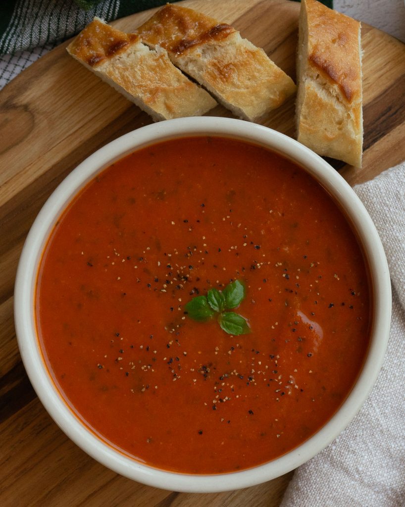 bowl of roasted tomato basil soup with on a wooden cutting board with 3 pieces of baguette bread 