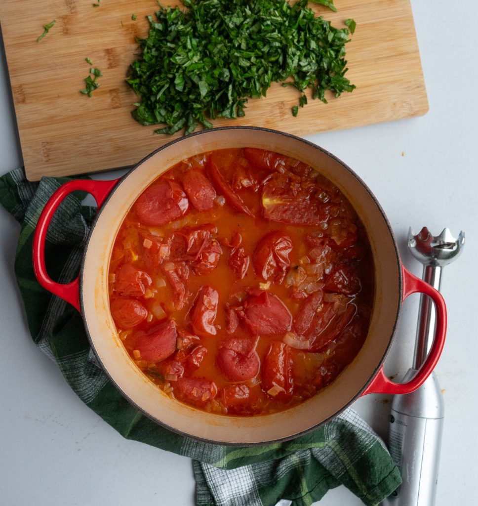 dutch oven with roasted tomatoes in it, chopped basil is on a wooden cutting board to the top of photo, immersion blender is on the side, a green plaid kitchen towel is under the dutch oven