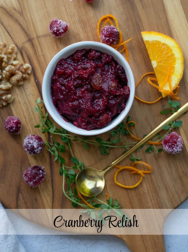 cranberry relish in a dish on a wood cutting board surrounded by the relish ingredients, with a gold spoon laying to the bottom right of the bowl