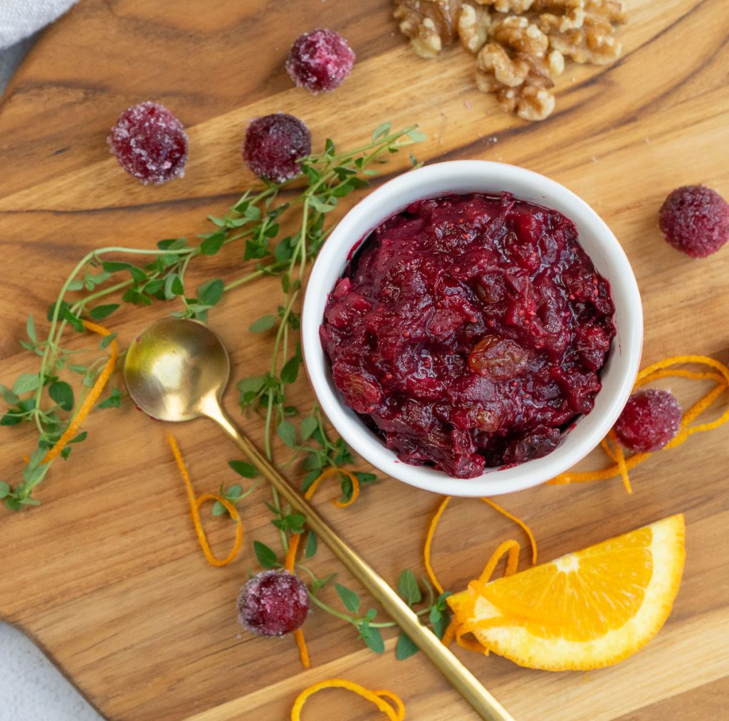 dish of cranberry relish on a wood cutting board surrounded by cranberries, an orange slice and some fresh herbs a gold spoon is laying off to the left side of the dish