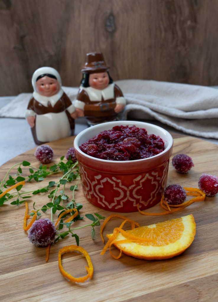 dish full of cranberry relish with spices, orange zest scattered around the dish, pilgrim salt and pepper shakers are in the background