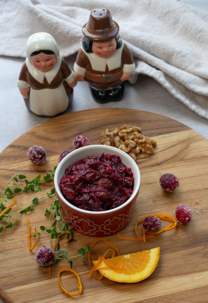 cranberry sauce on a wood cutting board with ingredients encircling the dish, pilgrim salt and pepper shaker in backrground