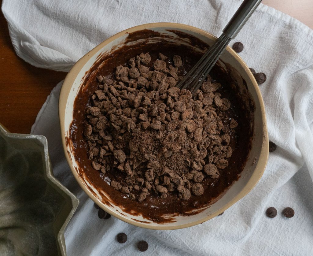 chocolate chips on top of chocolate cake batter in a glass bowl with whisk sticking out the top right of bowl. bowl is surrounded by a white cloth sprinkled with chocolate chips