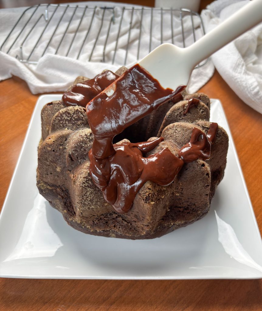 spooning chocolate ganache over triple chocolate bundt cake . cake is on a white cake plate 