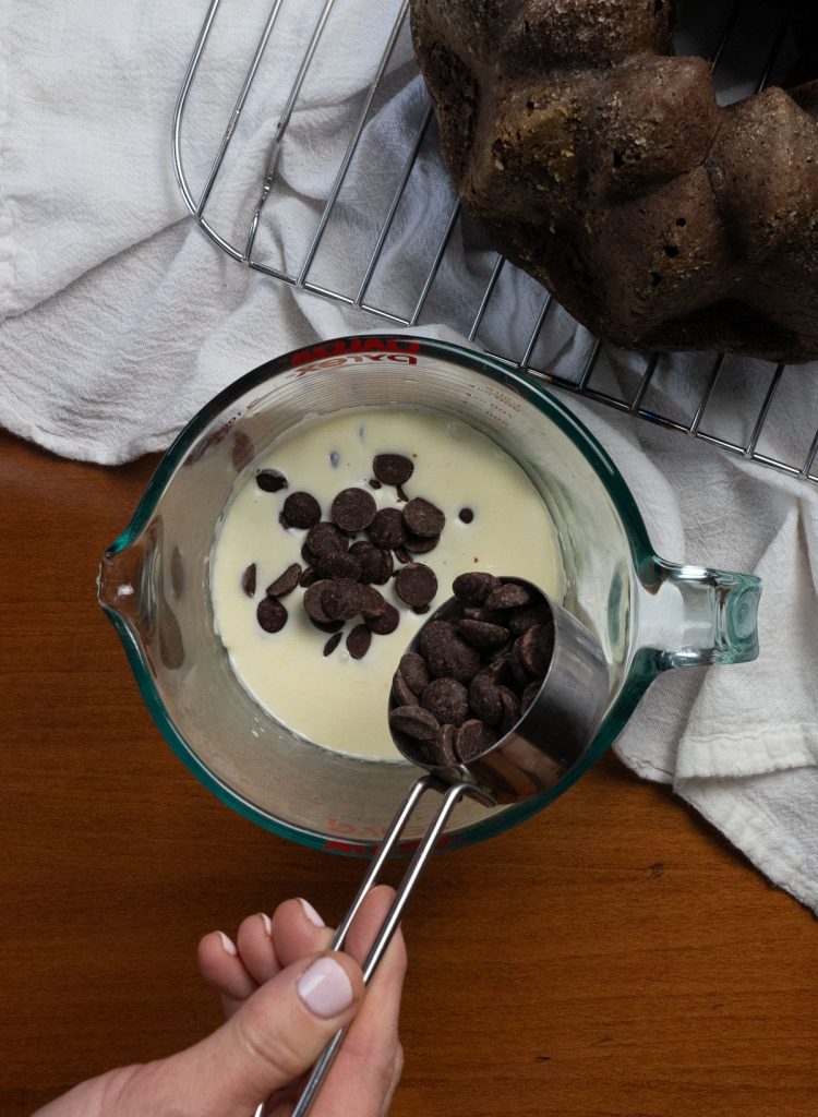 making chocolate ganache for the triple chocolate bundt cake measuring cup with heavy cream, hand pouring in chocolate chips to cream