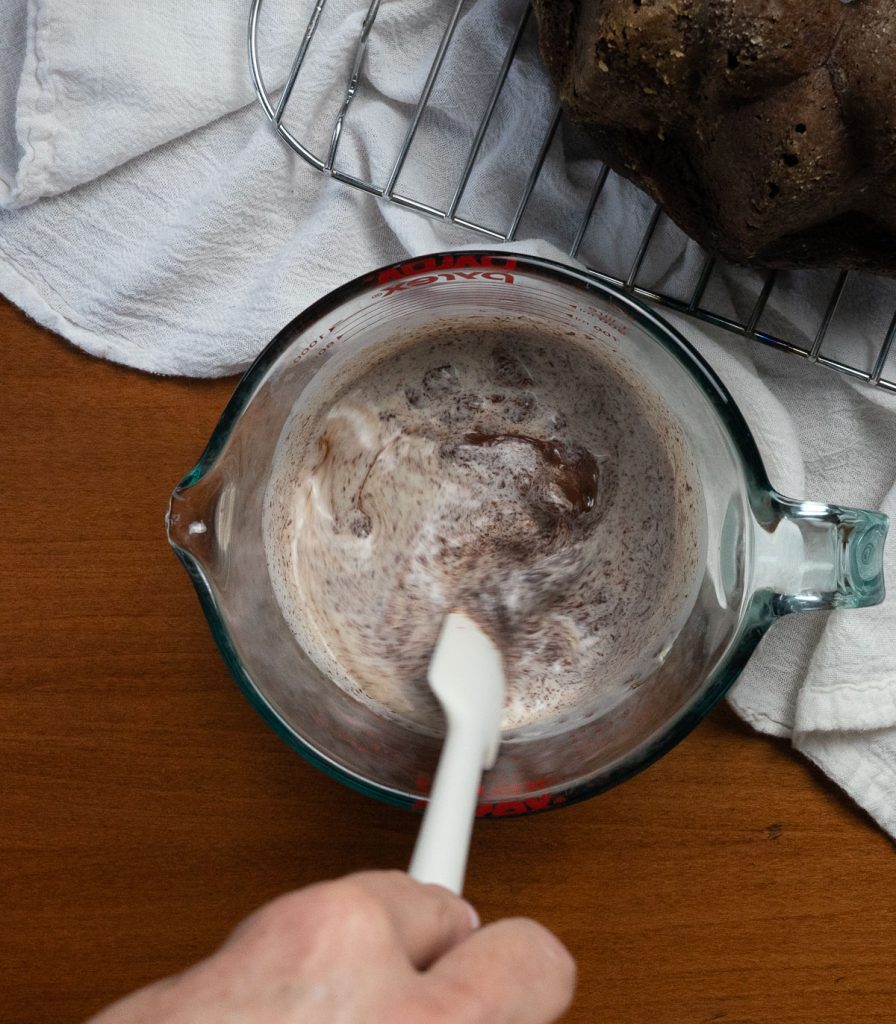 stirring chocolate chips into hot cream with a white spatula on a wood surface, cake is in the upper right corner of the photo