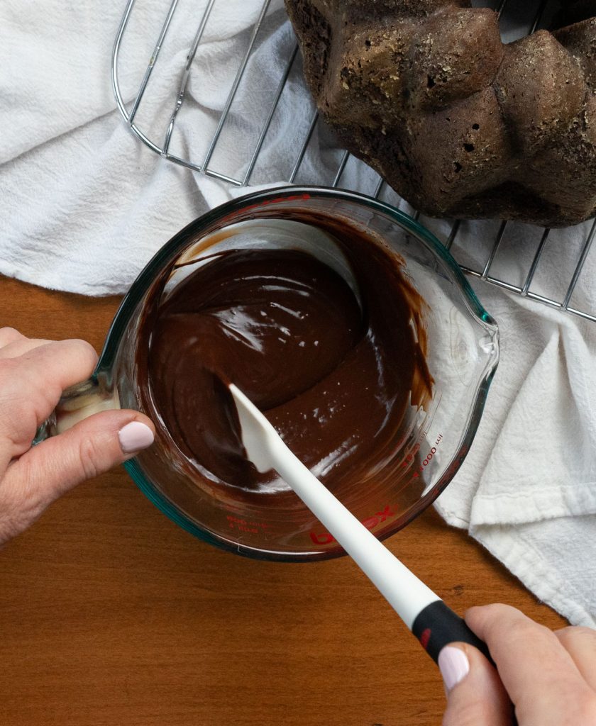 white spatula stirring chocolate ganache in a large glass measuring bowl, cake is in upper rt corner of the photo on a cooling rack