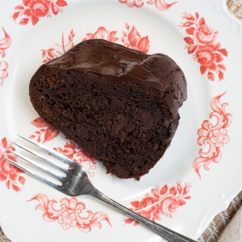 one piece of dark chocolate cake on a white plate with floral design around the edge and a fork laying across the bottom