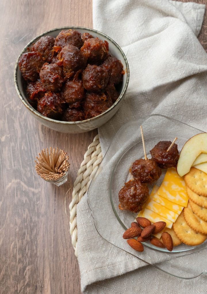 bowl of meatballs with cocktail toothpicks in front of it, on a wooden countertop. small glass plate of appetizer in front off to the right side of appetizer meatballs