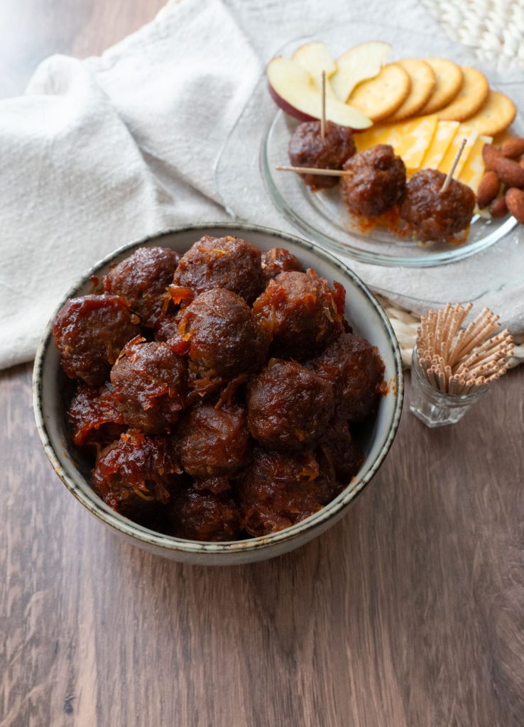 sweet and sour appetizer meatballs in a tan bowl on a wood countertop with a plate of meatballs,  crackers and cheese, nuts, and apples in background