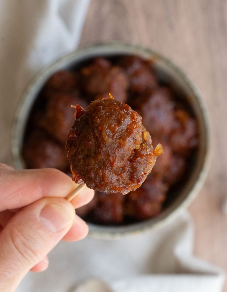 close up of a hand with a meatball on a toothpick with a bowl of meatballs in the background on a wood countertop 