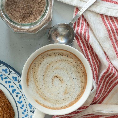 cup of mocha cappuccino next to a cookie on a plate, jar of the cappuccino mix is in upper left corner with a measuring spoon