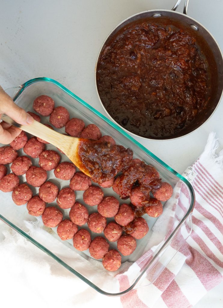 glass dish lined with meatballs, sauce in sauce pan , wooden spoon is used to spoon sauce over the meatballs