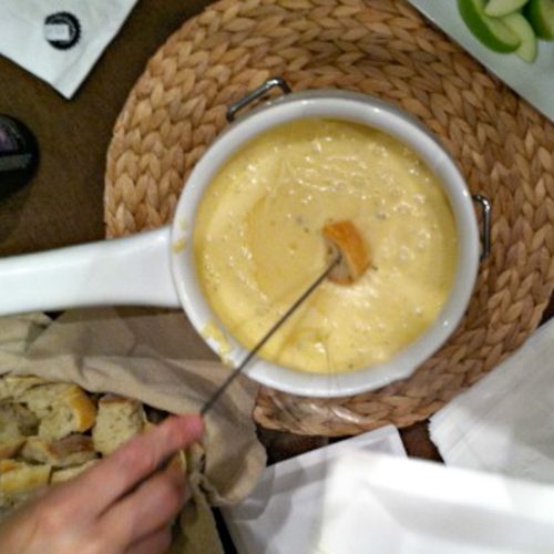 fondue pot in the middle of photo with apple slices in upper right hand corner and bread cubes in lower left a hand with a fondue fork dipping bread into melted cheese fondue