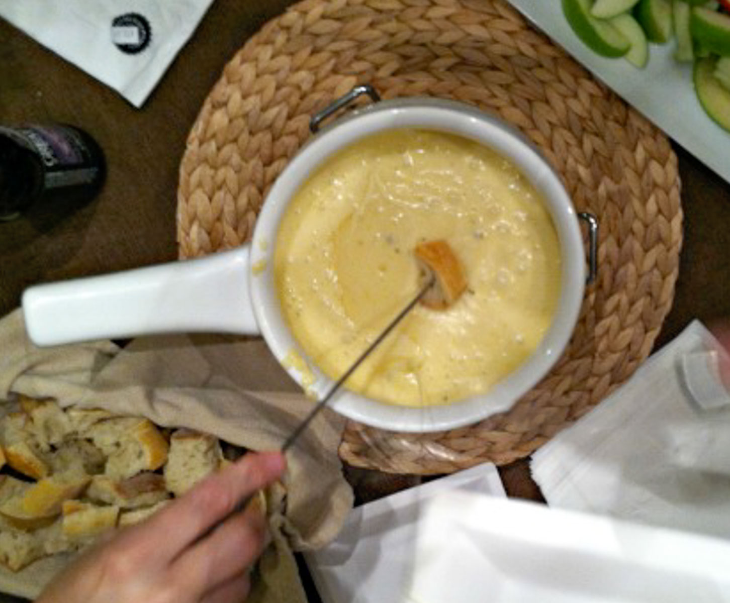 fondue pot in the middle of photo with apple slices in upper right hand corner and bread cubes in lower left a hand with a fondue fork dipping bread into melted cheese fondue