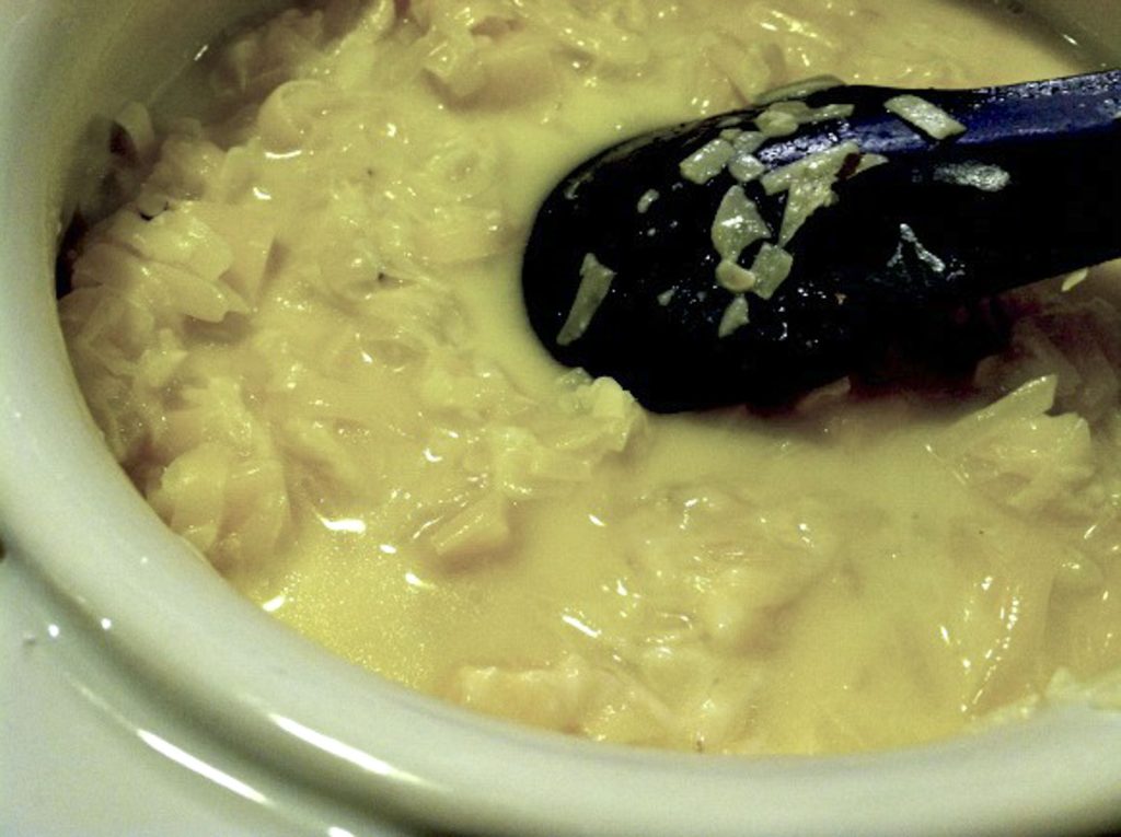 Shredded cheese in fondue pot starting to melt, but also still clumpy. Black spatula is in the pot stirring the cheese. 