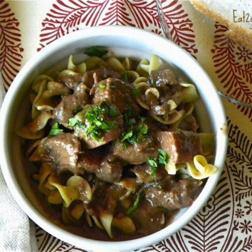 beef stew over wide egg noodles in a round white bowl on a place matt