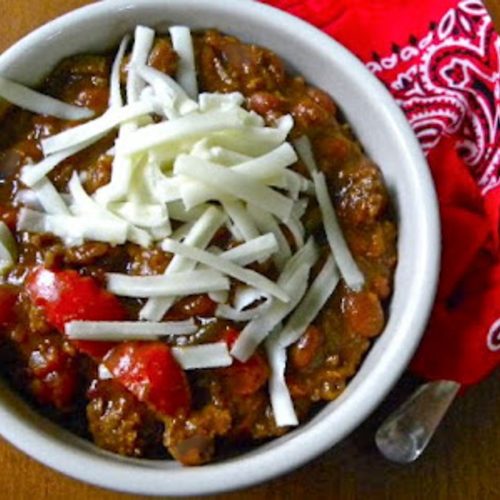 chili in a white bowl with white monterey jack cheese shredded on top, bowl is sitting on a red bandana with a silver spoon to the right of the bowl.