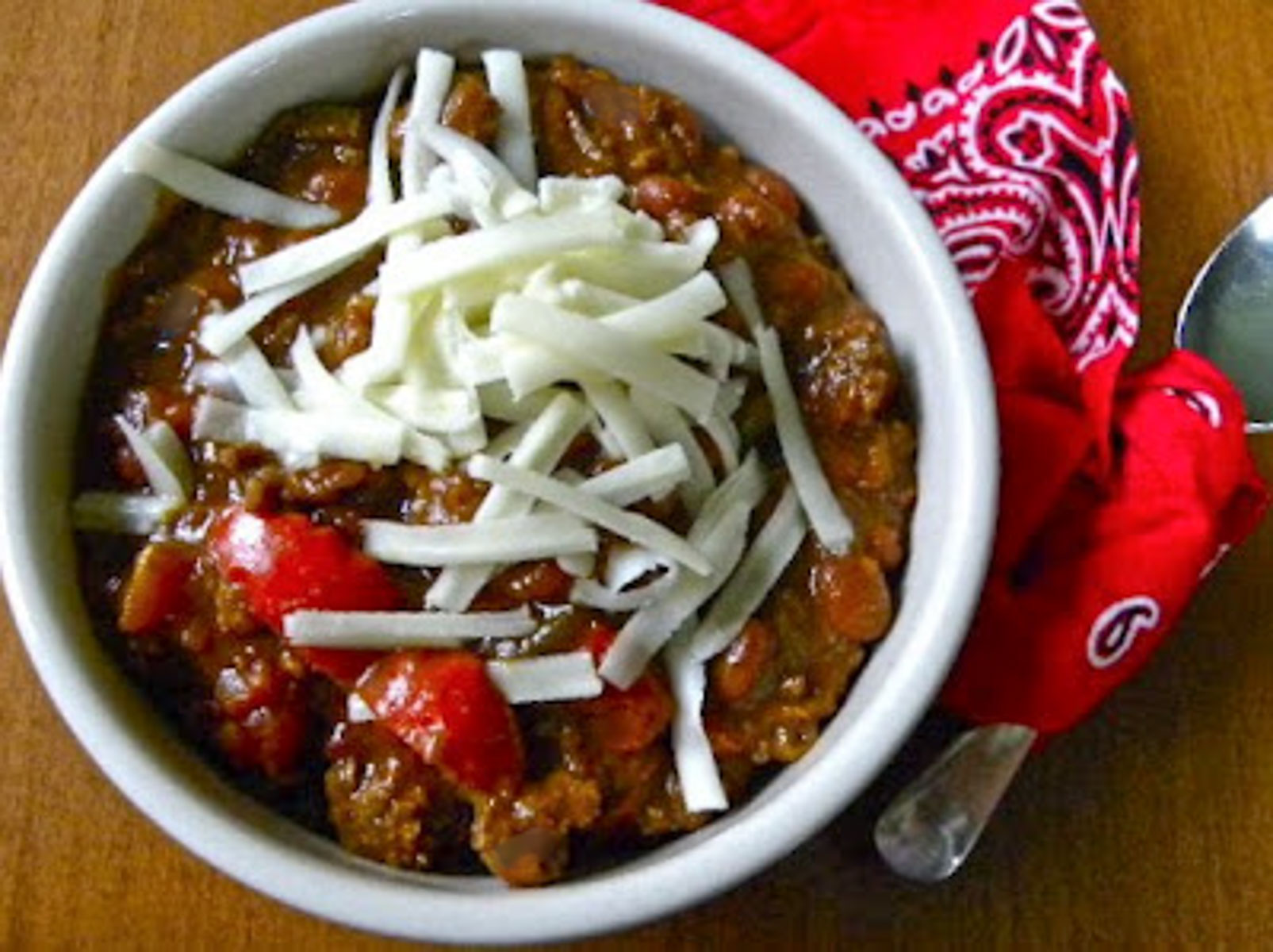 chili in a white bowl with white monterey jack cheese shredded on top, bowl is sitting on a red bandana with a silver spoon to the right of the bowl.