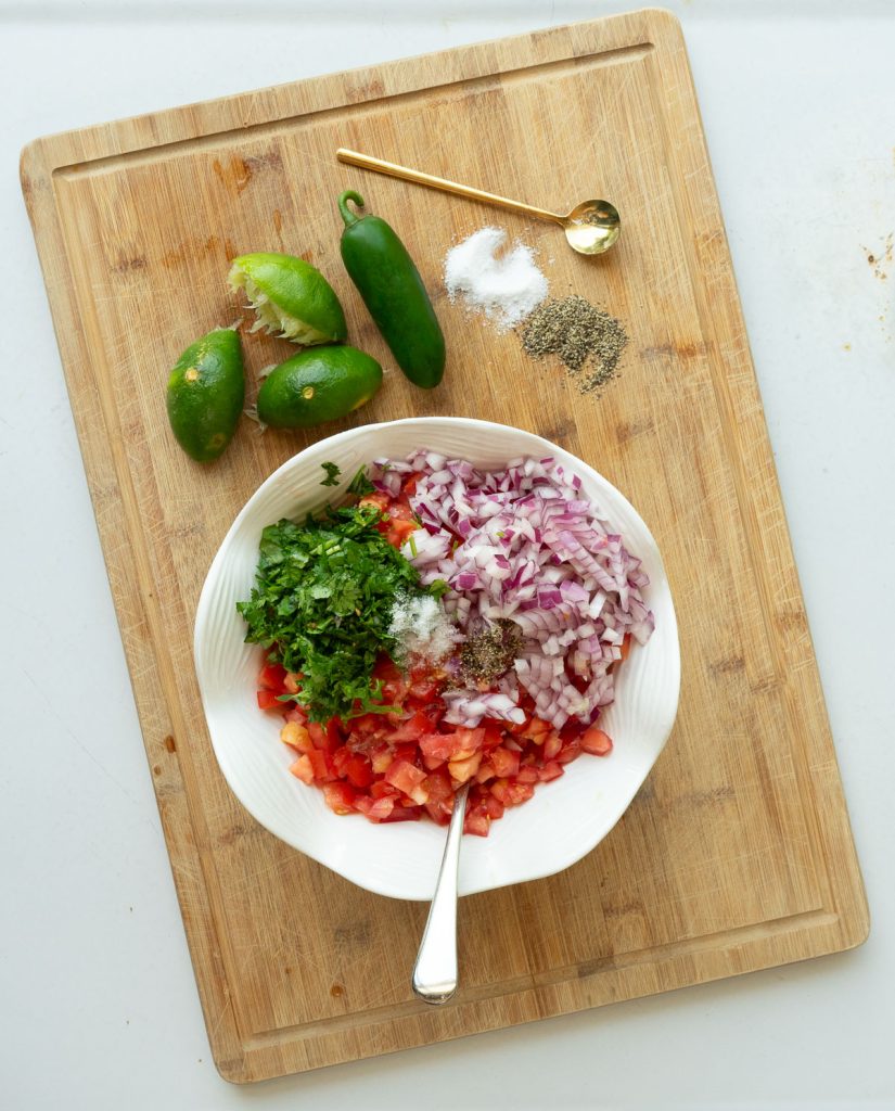 white bowl with chopped tomato, onion, cilantro, salt and pepper sitting on top of a wood cutting board there is a half squeezed lime some salt and pepper and a jalapeno on the cutting board with a gold spoon