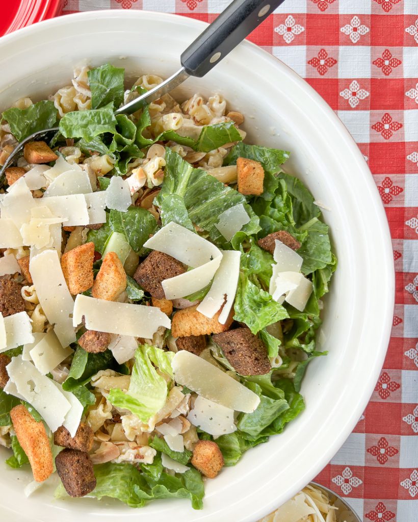 close up of bowl of  chicken caesar pasta salad with parmesan spoon is in the bowl in top of picture, bowl is sitting on a red and white checkered tablecloth 