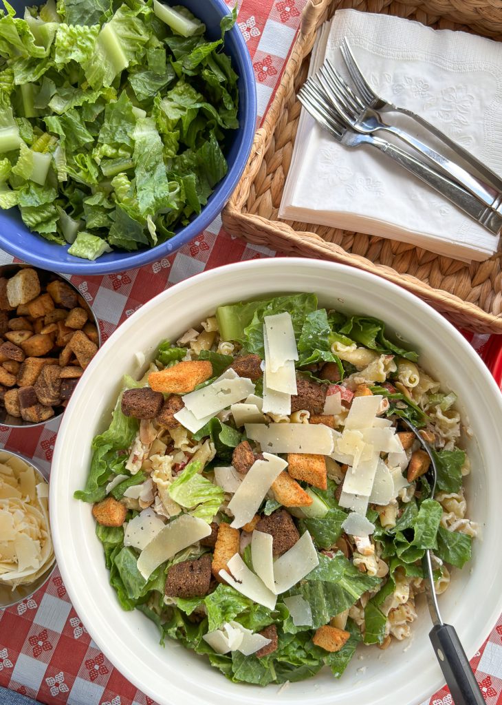 large bowl in bottom half of photo has chicken caesar pasta salad in it, side dishes of parmesan and croutons to the left, blue bowl full of romaine lettuce, square wicker basket in upper right hand corner with napkins and forksss all on a red and white checkered tablecloth