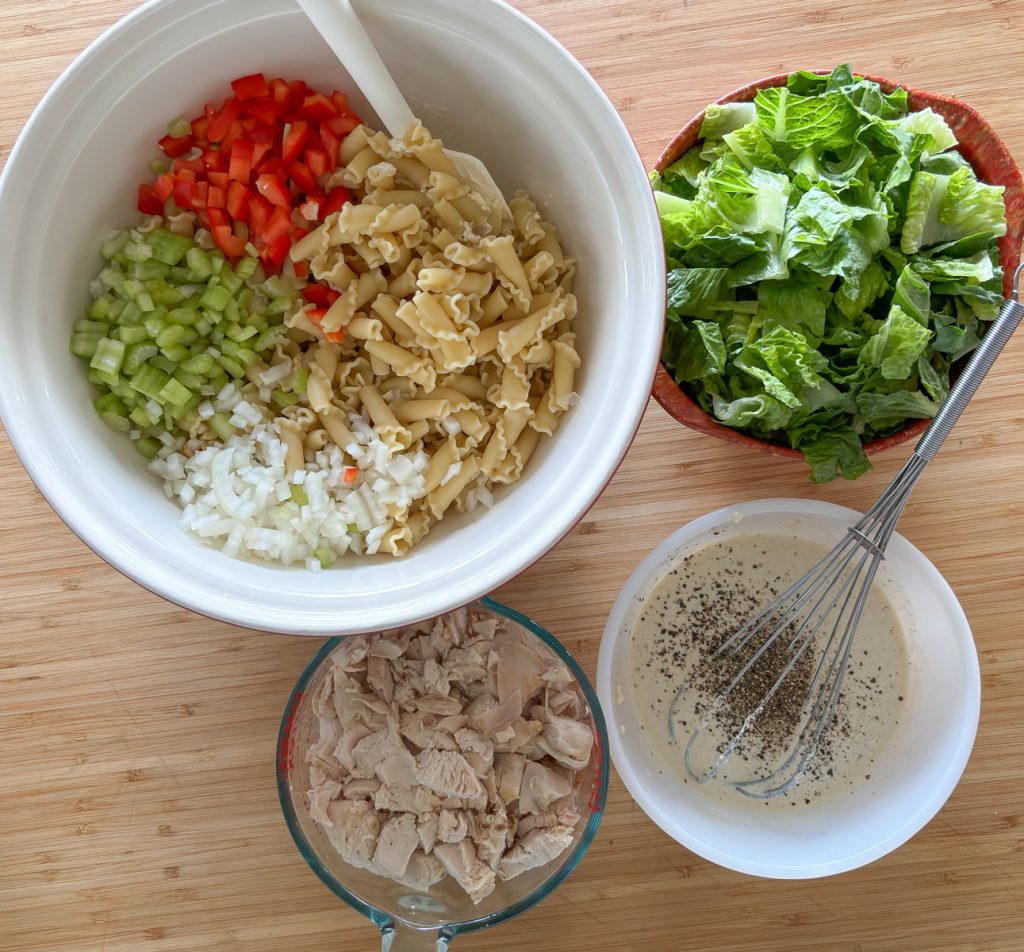 bowl with pasta, red bell pepper, onion, and celery. counter clockwise bowl with chunks of chicken, dressing for salad, bowl of romaine lettuce