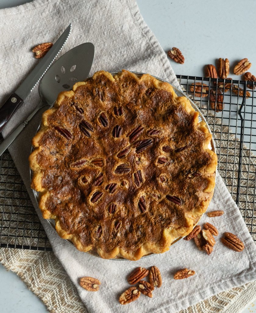 whole uncut pecan pie on a tan cloth napkin with pie server and knife on upper left side of photo