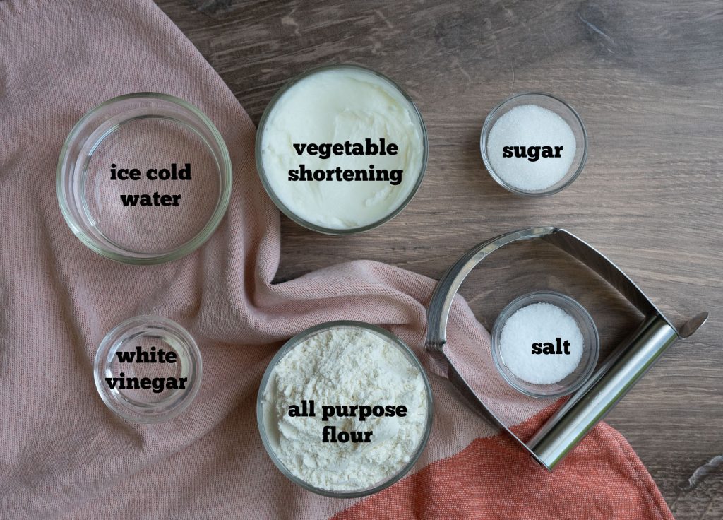 ingredients for pie crust on a wood cutting board with a peach/orange colored dish towel  and a pastry cutter 