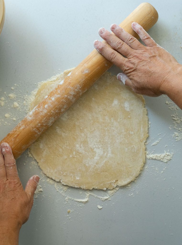 rolling out pie dough into a circle with a wood rolling pin on a gray countertop