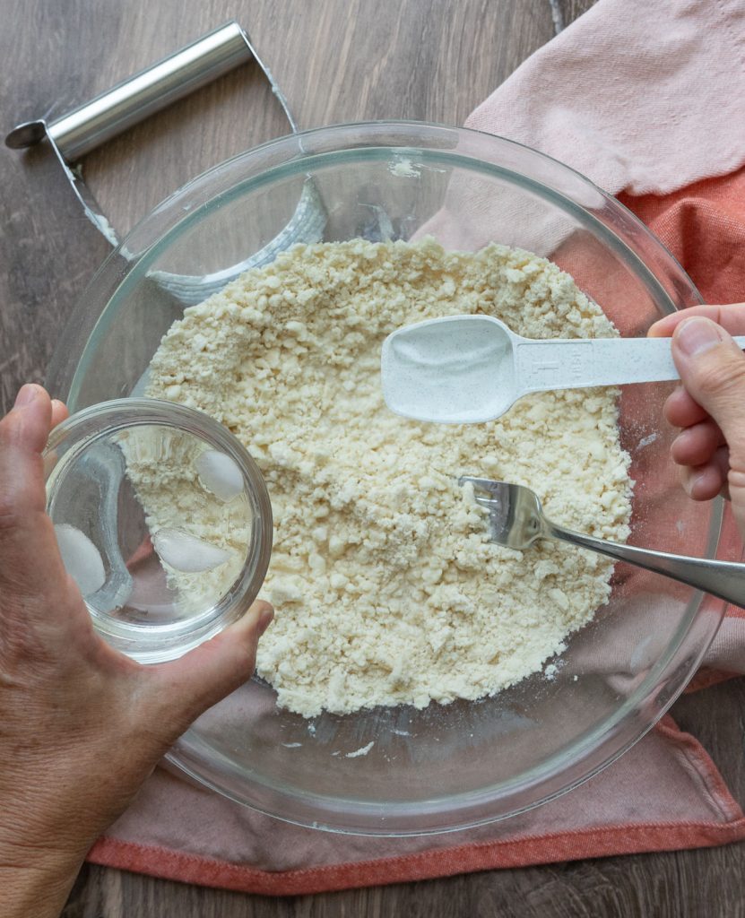 drizzling ice water with vinegar over the dry ingredients for pie crust in a bowl. bowl is setting on wood surface and a rust colored towel