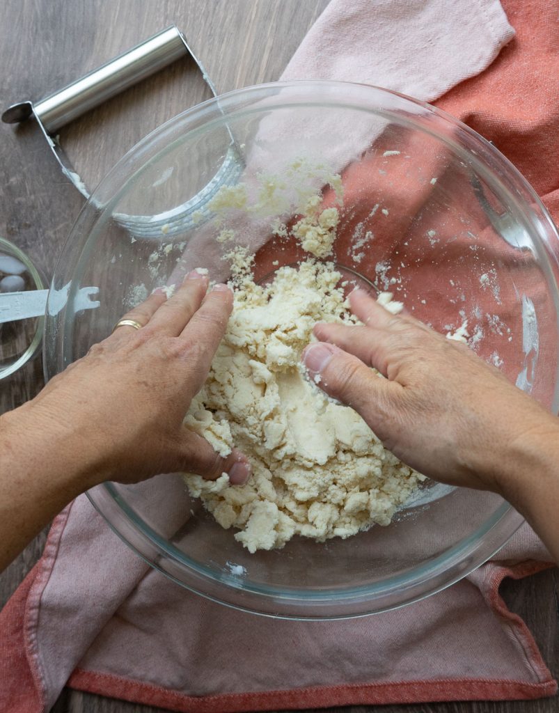 glass bowl with hands gathering crust together into a ball, on a wood surface with rust colored kitchen towel and a pastry cutter in background