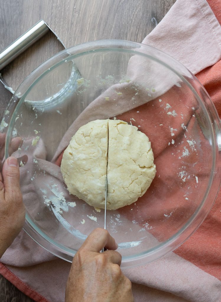 ball of pie dough in bottom of a glass bowl on a wood surface with a rust colored kitchen towel and a pastry cutter in the background.  hand is cutting the pie crust in half