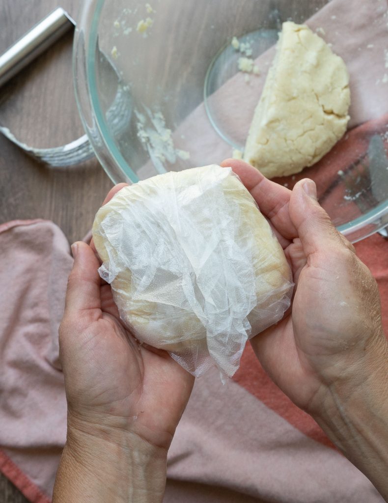 holding a plastic wrapped pie crust in two hands glass bowl with pie dough in it, a pastry cutter and a rust colored kitchen towel are all in the background