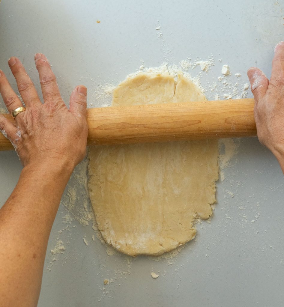 rolling out pie dough out with a wood rolling pinon a lightly floured gray countertop 