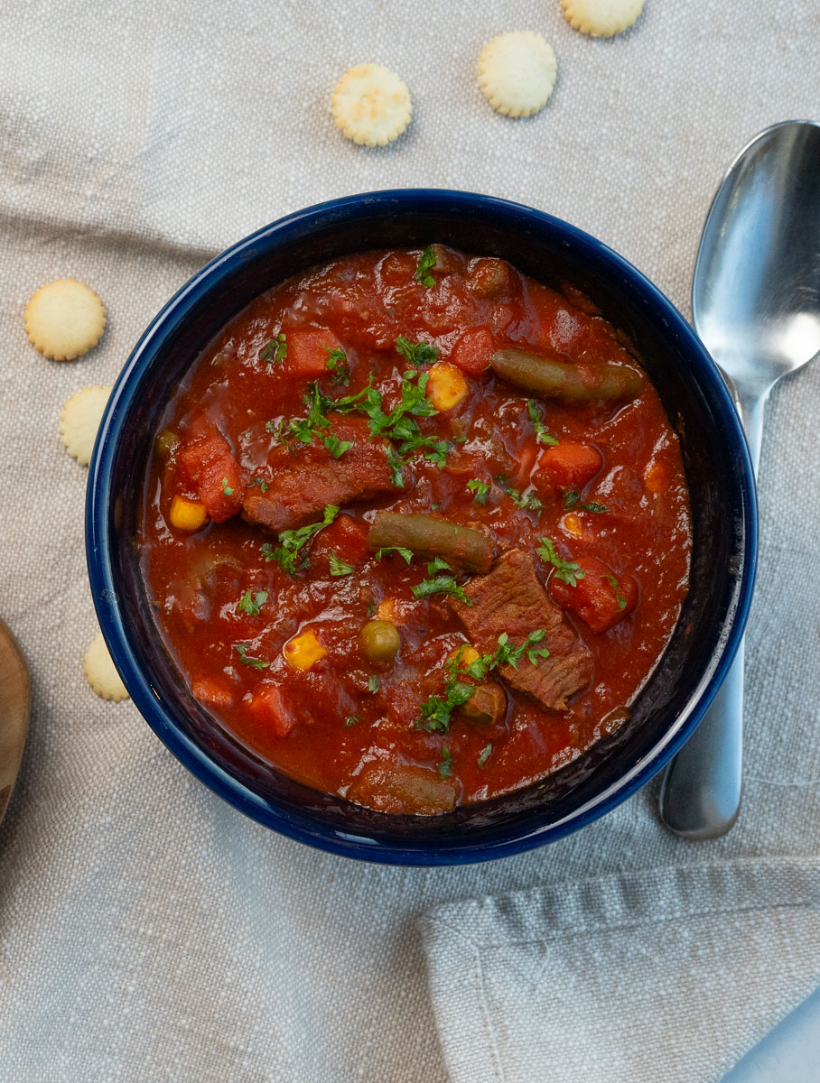 blue soup bowl full of hearty steak soup spoon to the right of the bowl, sup crackers strewn around on ta ble