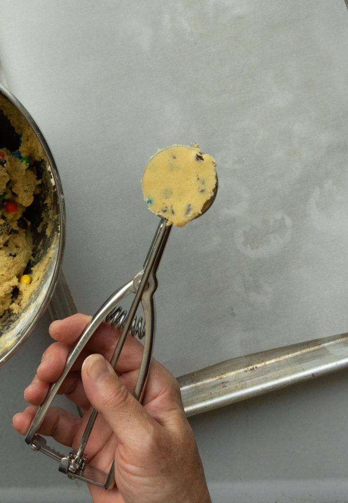 cookie scoop full of cookie dough with a baking sheet lined with parchment paper on counter top behind it