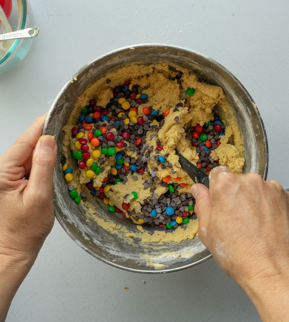 adding chocolate chips and m&m's to cookie dough in a silver bowl on a gray countertop