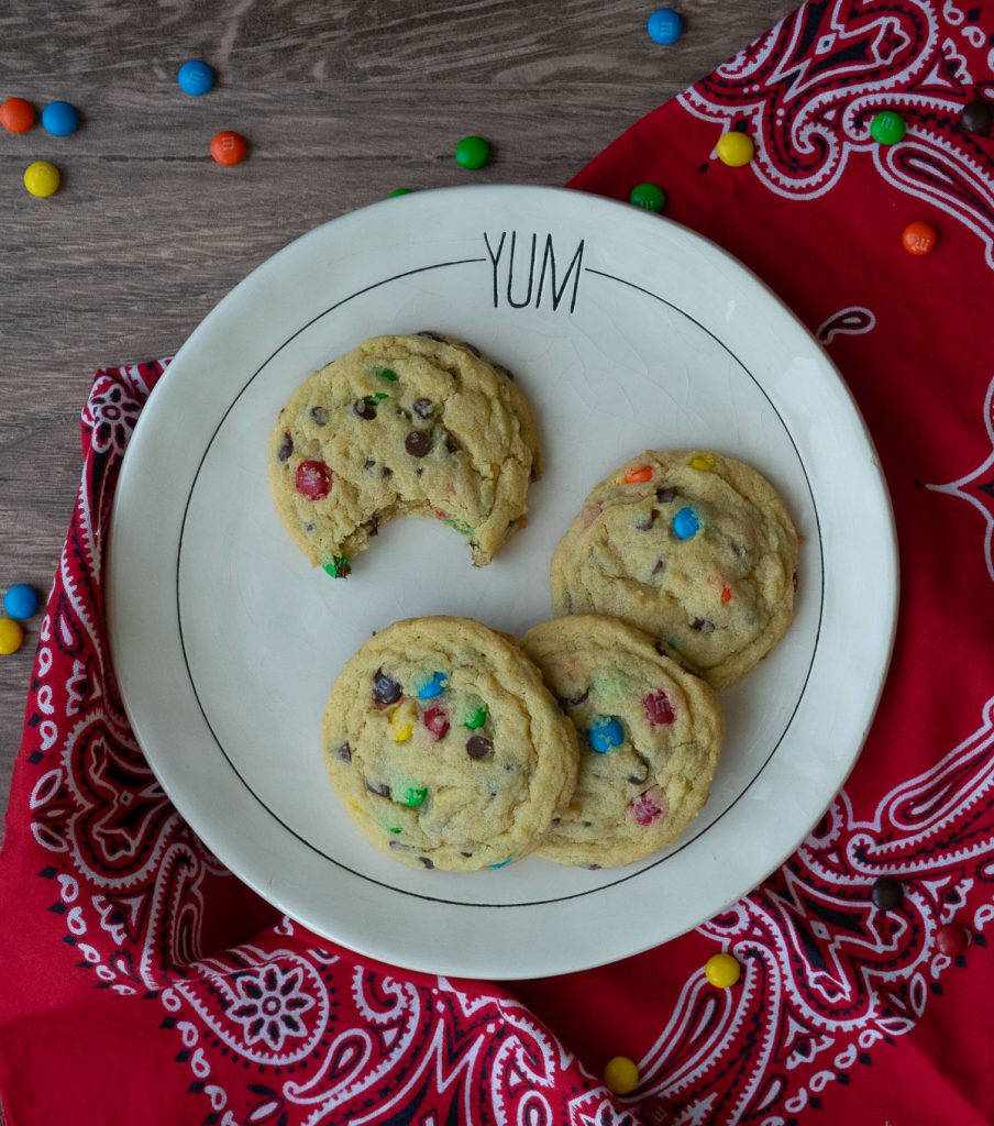 four M&M chocolate chip cookies on a white plate that says YUM at the top one of the cookies has a bite out of it, m&m's are scattered on the table and the plate is sitting on a red bandana
