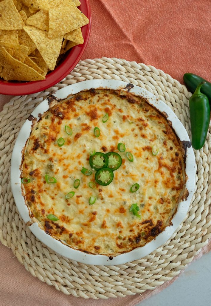 white round dish of jalapeno cheesy chicken dip on a rattan place mat with a bowl of tortilla chips in upper right corner