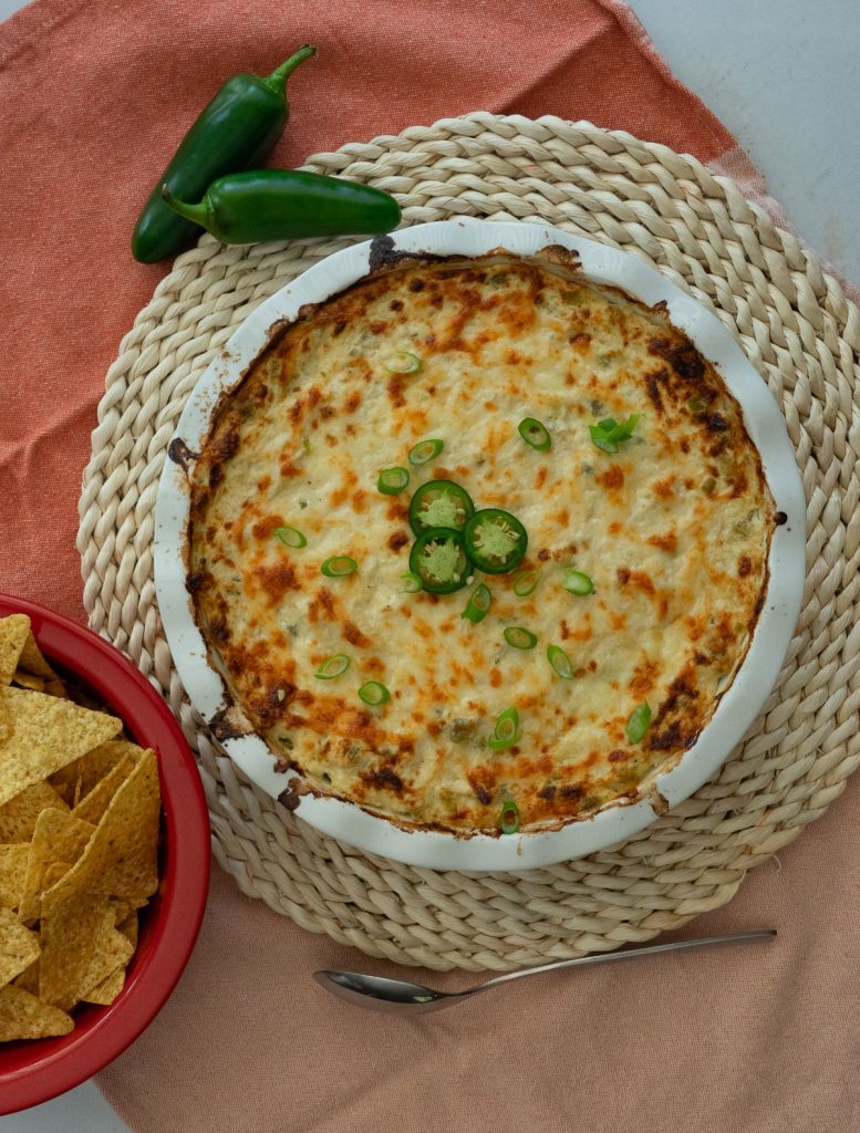 white round dish of jalapeno cheesy chicken dip on a rattan place mat with a bowl of tortilla chips in upper right corner