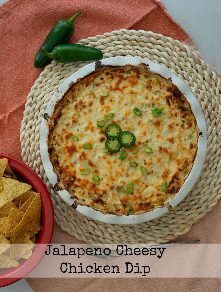 white round dish of jalapeno cheesy chicken dip on a rattan place mat with a bowl of tortilla chips in upper right corner