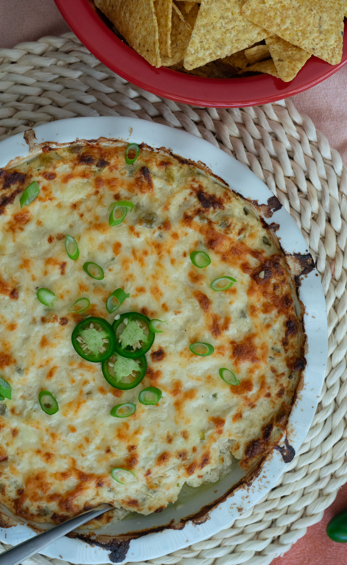 white round dish of jalapeno cheesy chicken dip on a rattan place mat with a bowl of tortilla chips in upper right corner