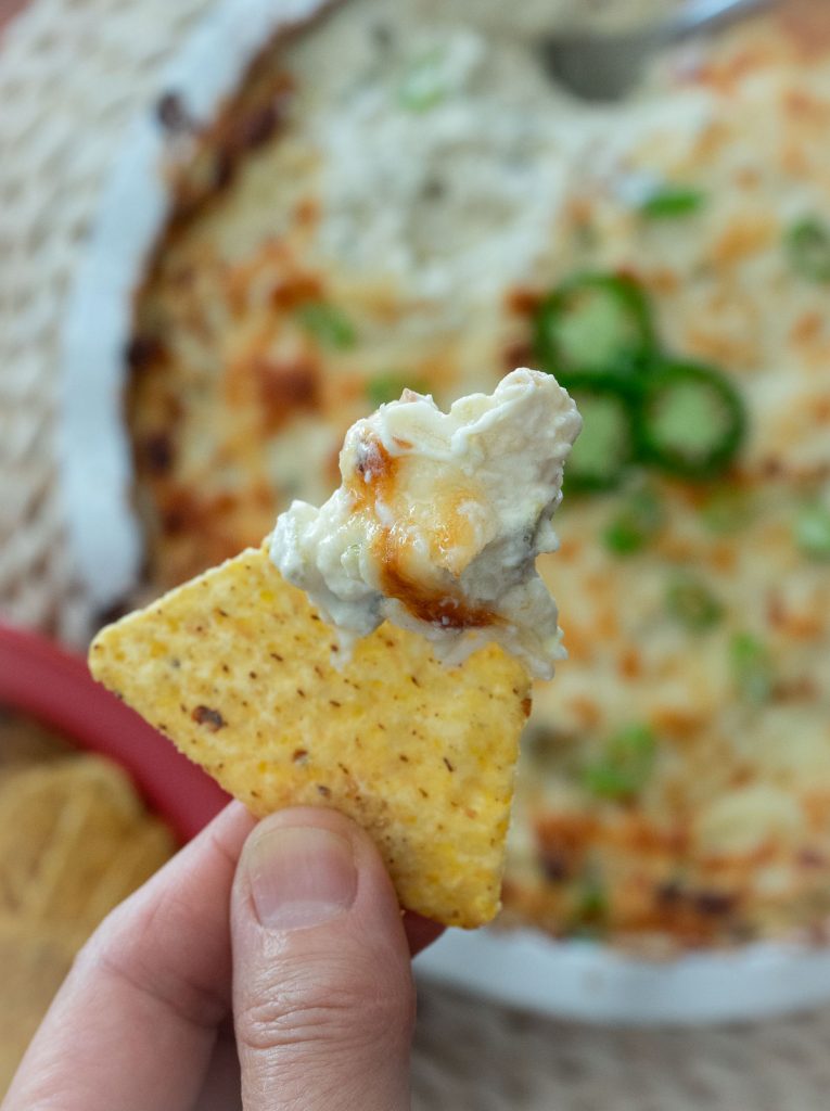 chip with jalapeno cheesy chicken dip on it held over a dish of baked dip