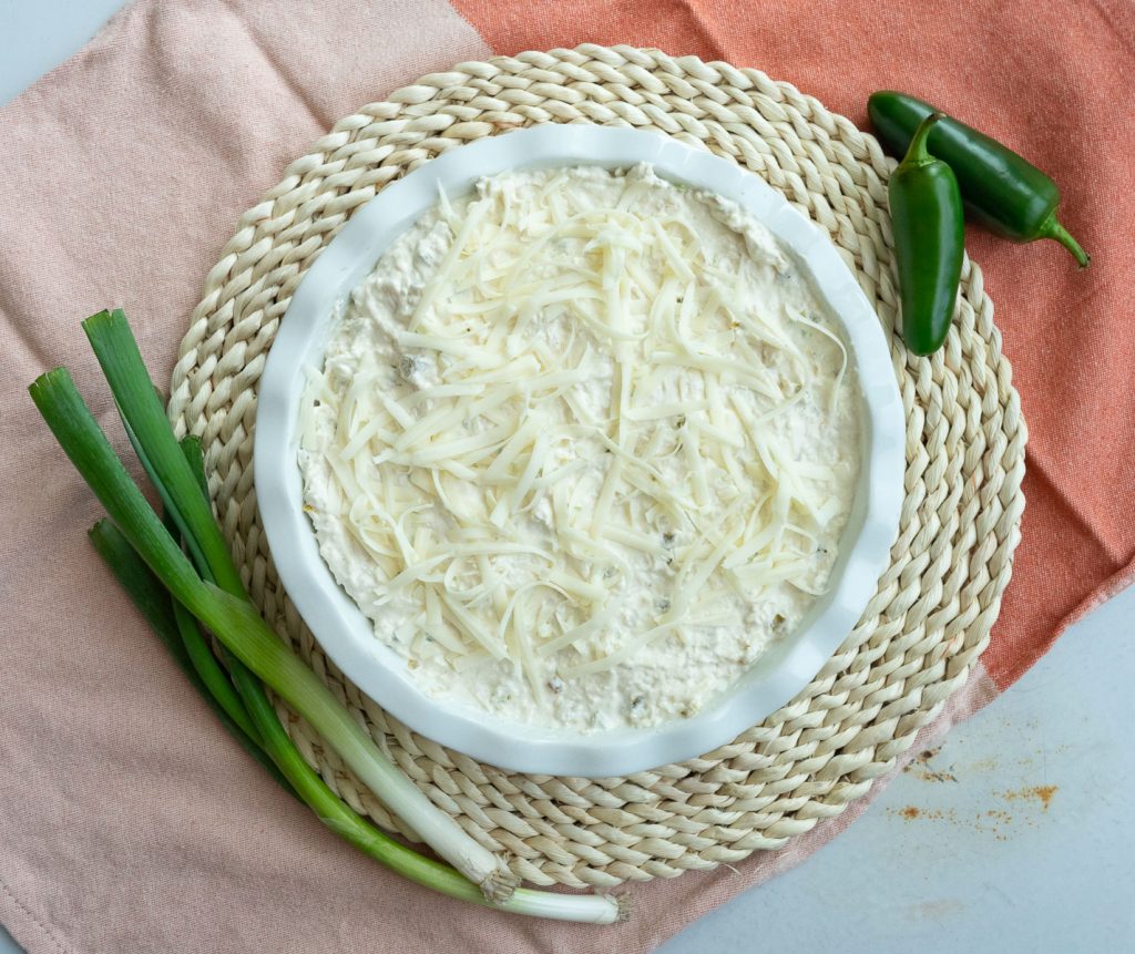 unbaked jalapeno cheesy chicken dip in a white round baking dish with green onions on the lower left side of the dish and jalapeno peppers on the upper right side of the dish