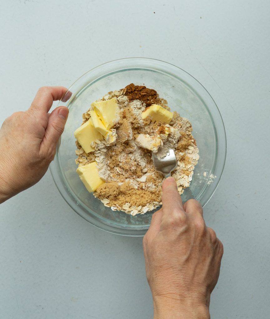 butter, flour, brown sugar, oatmeal and cinnamon in a glass bowl being combined with a fork