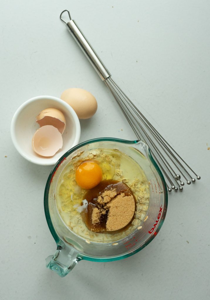 eggs, oil, brown sugar in large glass mixing bowl on a gray table whisk and small white bowl with eggs shells in it